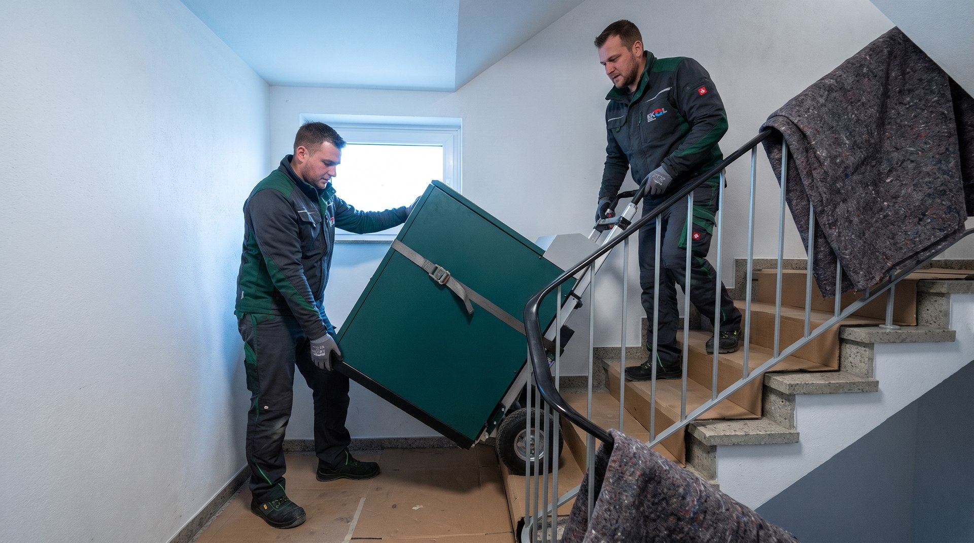 Zwei EKOL-Mitarbeiter transportieren ein SenerTec Dachs mit einer Treppensteigkarre in den Keller